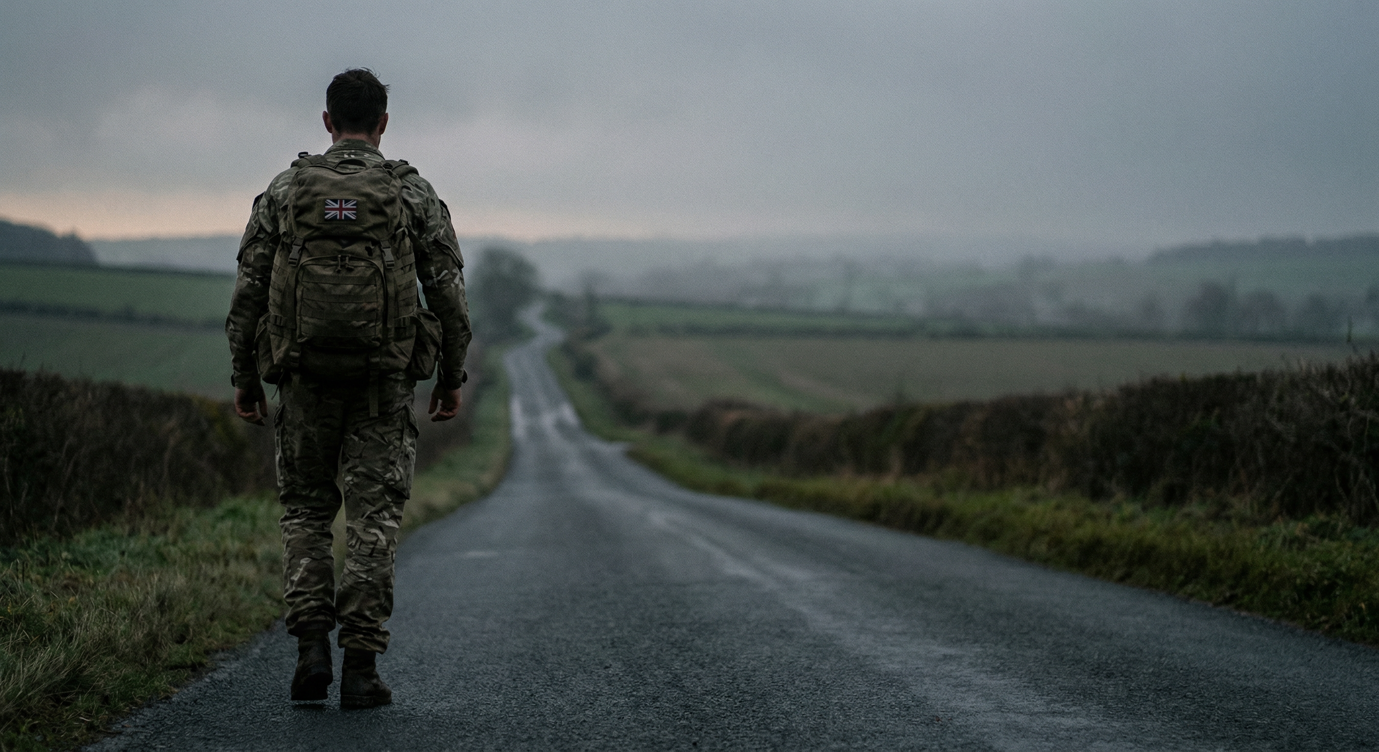 British soldier walking alone down a misty rural road in full combat gear, seen from behind